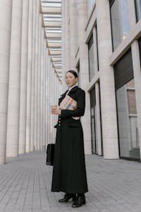 Stylish businesswoman in formal attire holding coffee and newspaper outdoors in an urban setting.