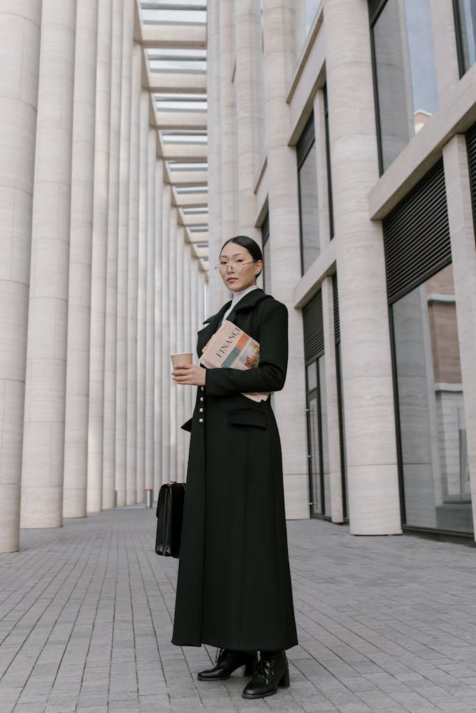 Stylish businesswoman in formal attire holding coffee and newspaper outdoors in an urban setting.