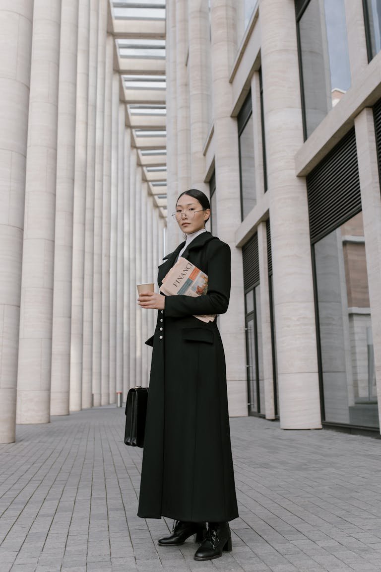 Stylish businesswoman in formal attire holding coffee and newspaper outdoors in an urban setting.