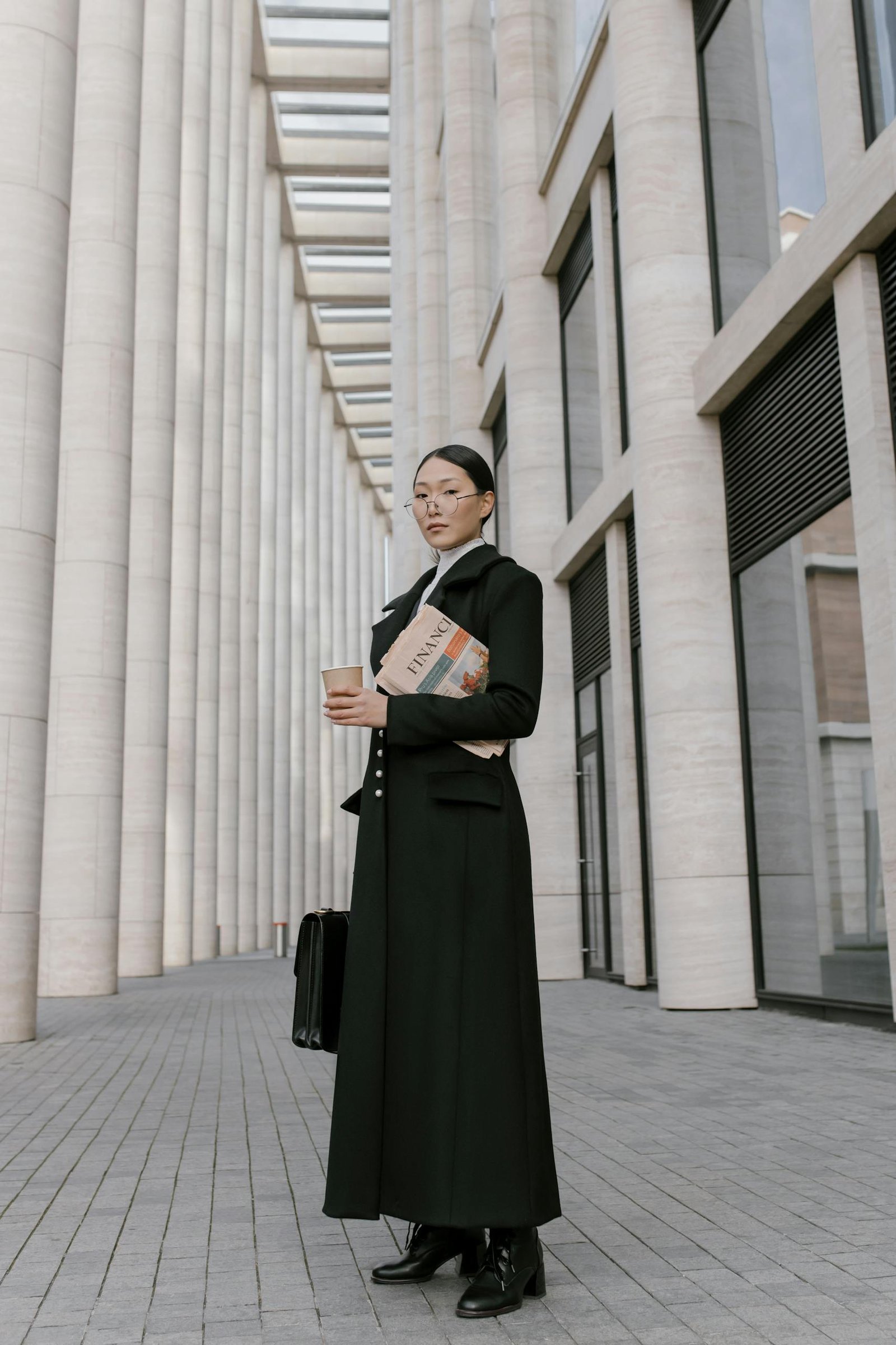 Stylish businesswoman in formal attire holding coffee and newspaper outdoors in an urban setting.